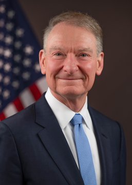 An official portrait of an older man in a dark suit and blue tie, standing in front of an American flag.
