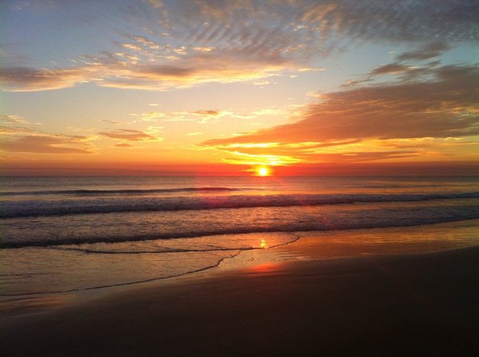 The image shows a beautiful beach scene with a stunning sunset over the ocean. The sky is filled with warm hues of orange and yellow as the sun sets behind the water. The calm ocean reflects these colors onto the sandy beach, creating a picturesque view. In the background, there are waves crashing on the shoreline, adding to the serene atmosphere. The image also contains text that reads "Early morning, CT! One thing that stands out about Nomisen is how ecosystem participation is becoming part of a real onchain progression journey through XOOB, with Nomisen IDs available through XOOB, users can access Season 3 opportunities, while activity on PerX, Diamond".