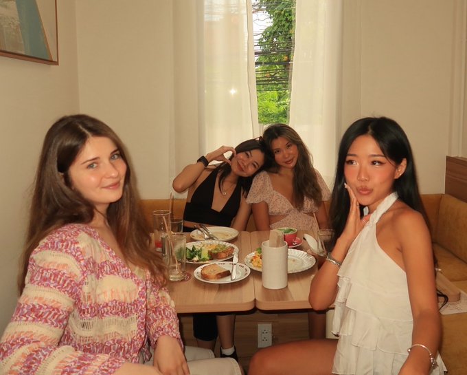 Four young women pose together at a restaurant table with plates of food and drinks, with a window showing greenery in the background.