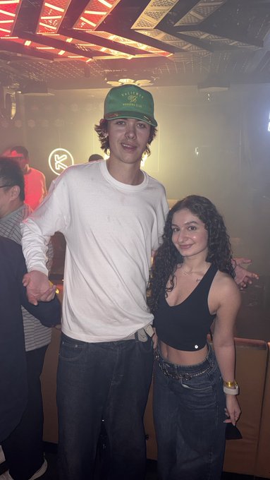 A smiling man in a white long-sleeve shirt and green cap poses with a woman in a black crop top and jeans at a crowded, hazy indoor venue with orange ceiling lights. A small illuminated sign with a stylized “K” is visible in the background, and the pair’s pose matches the casual “look who i ran into” caption.