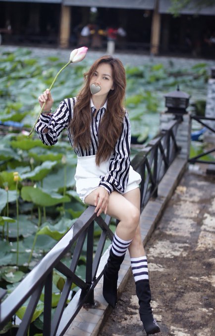 A woman in a black and white striped shirt and white shorts sits on a fence by a lotus pond, holding a pink lotus flower near her face.