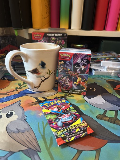 A bird-patterned mug filled with coffee next to Pokémon Trading Card Game products, including a Destined Rivals Booster Bundle box and a Mega Evolution booster pack, displayed on a colorful playmat.
