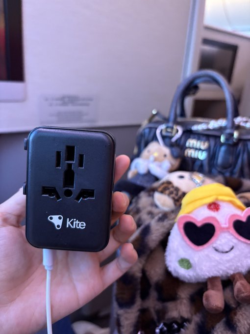 A hand holds a black "Kite" branded universal travel adapter with a white charging cable plugged in, shown against the backdrop of a Miu Miu bag and a plush doll wearing heart-shaped sunglasses. The image ties to the post about traveling to Korea for ICML, showcasing GoKite AI swag as part of the travel essentials for the trip.