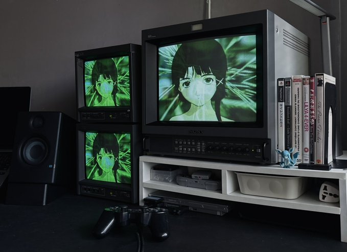 Retro gaming setup featuring multiple CRT monitors displaying an anime-style character, a Sony monitor on top of a console, a speaker on the left, and a row of retro game cases and books on the right.