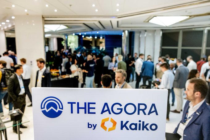 A conference networking event features a large sign reading “THE AGORA by Kaiko” in front of a crowded hall.