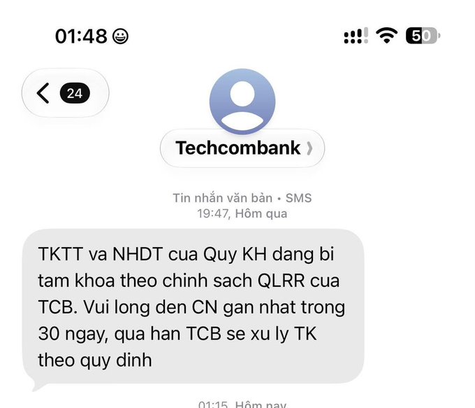 A smartphone screenshot of a Techcombank SMS notification in Vietnamese stating that the customer's payment account and savings account are temporarily locked per bank policy, requesting a branch visit within 30 days.
