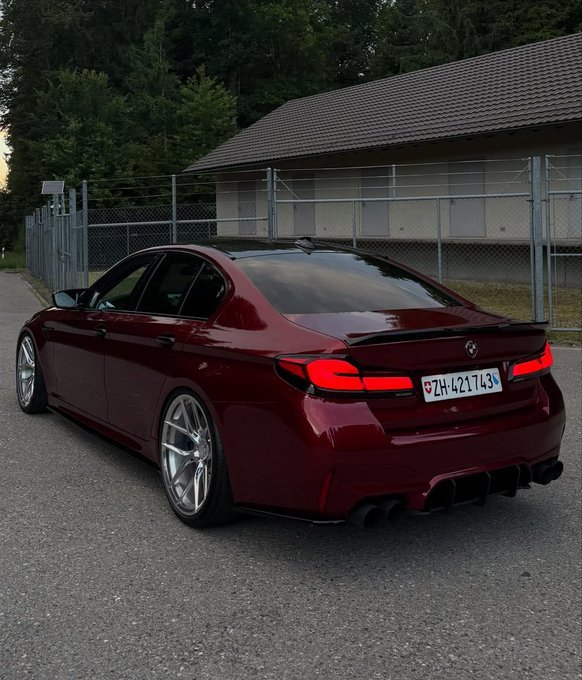 A dark red BMW sedan with custom wheels and a rear spoiler is parked on an asphalt surface next to a building with a fence.