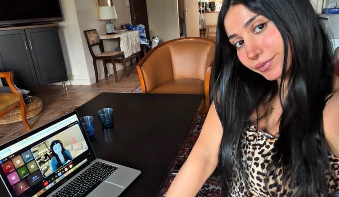 A young woman with long dark hair wearing a leopard print top takes a selfie in a living room, with an open MacBook on a black coffee table behind her.