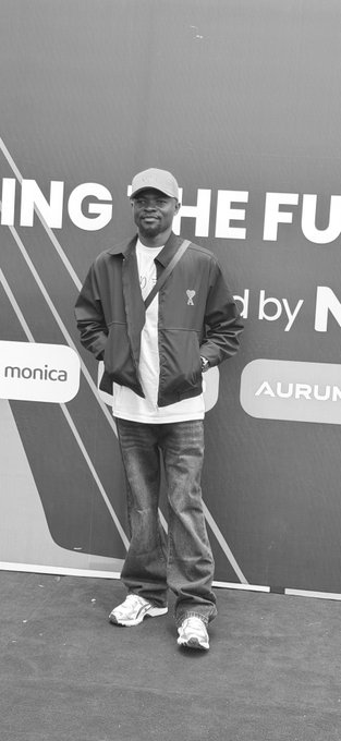 A smiling man in a cap, jacket, and jeans poses confidently in front of a sponsor backdrop reading "ING THE FU" and "by N," with "monica" and "AURUN" logos visible, capturing a moment from the NFT Unchain Summer event in Lagos.