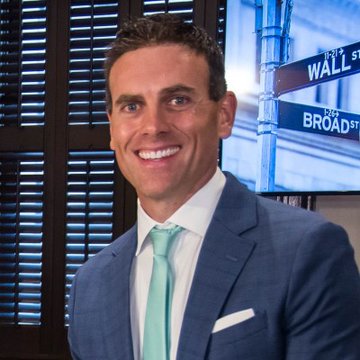 A professional headshot of a smiling man in a blue suit and light green tie, appearing to be in a TV studio with 'Wall Street' and 'Broad Street' street signs visible in the background.