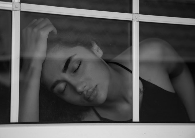 Black and white photograph of a woman with curly hair resting her head against a window pane, viewed through the divided glass with her eyes closed.