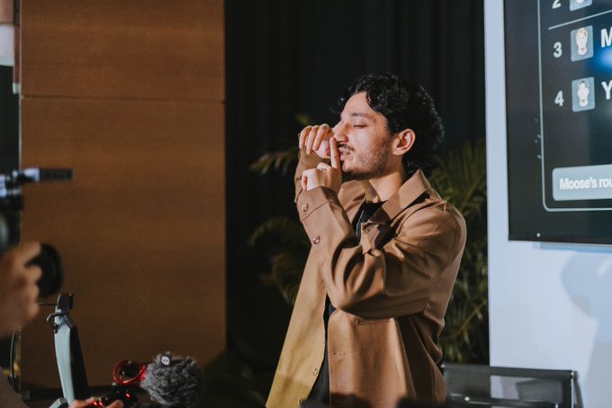 A man with dark hair and a beard wearing a tan jacket, standing on a stage with his hands clasped near his face, appearing emotional during what looks like a presentation or event.