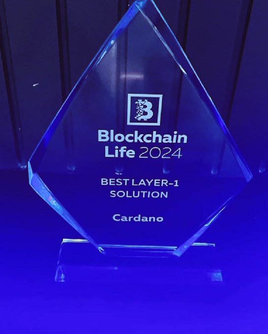 The image features a blue glass award, likely an award for best layer-j solution or something similar, placed on a table against a dark background. The award has a diamond shape with a white border, making it stand out prominently. The text "Best Layer-J Solution" is visible in the image, indicating that this award is related to a specific problem-solving technique or approach within the context of blockchain technology.