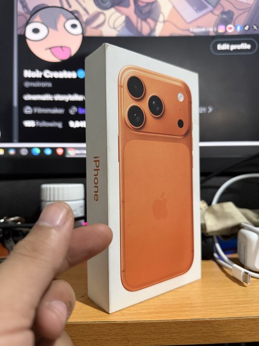 A hand holding an orange iPhone box (iPhone 17 Pro Max style) in front of a monitor showing a Noir Creates (@noironx) filmmaker profile page.