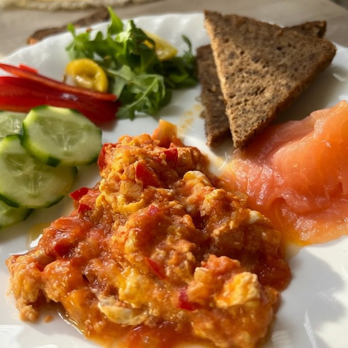 A plate of food featuring what appears to be shakshuka or scrambled eggs in tomato sauce, accompanied by smoked salmon, dark rye bread triangles, sliced cucumbers, red peppers, and a fresh green salad with lemon.