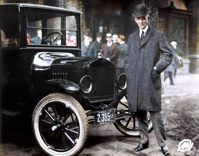 A colorized historical photograph of Henry Ford standing next to a black Model T Ford with a '2-385' Detroit license plate.