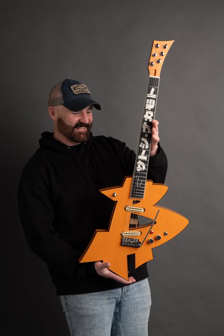 A bearded man wearing a black hoodie and a dark baseball cap smiles while holding up an orange, star-shaped electric guitar with 'HDRAHEAD' written on the fretboard.