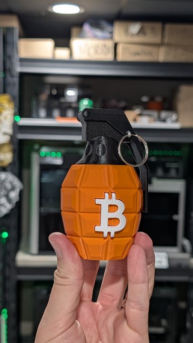 A hand holding an orange grenade-shaped Bitcoin keychain with the white Bitcoin 'B' logo against a background of shelves with boxes.