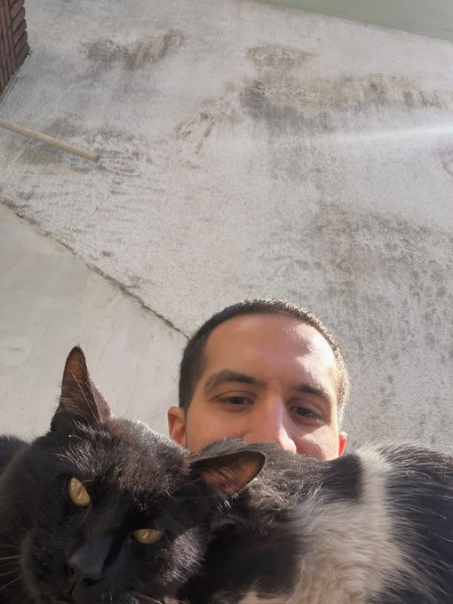 A man takes a selfie with a fluffy black and gray cat on a sunlit rooftop or balcony.