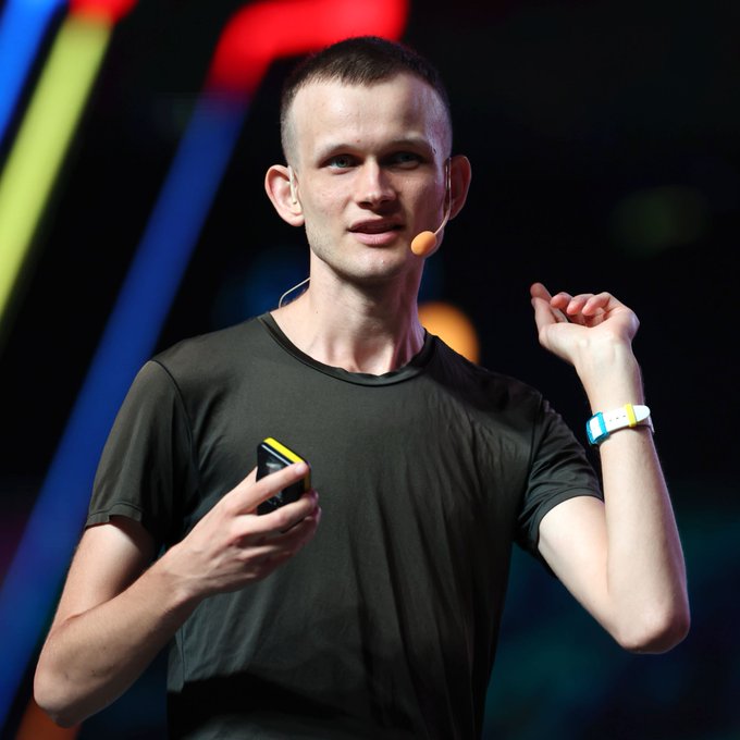 Vitalik Buterin, the co-founder of Ethereum, speaks on stage wearing a headset microphone while holding a presentation clicker. The caption relates to the post discussing Ethereum's Glamsterdam upgrade going live on the Sepolia testnet, with Buterin's presence tying to his role as the face of $ETH development.