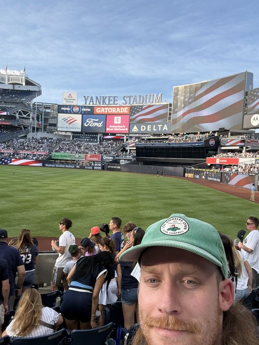 A man wearing a green "Shuffle" cap takes a selfie at Yankee Stadium, with the iconic "YANKEE STADIUM" sign and a giant American flag visible on the scoreboard behind him, visually confirming his lifelong dream of visiting the ballpark that's tied to his mention of #thanksshuffle.