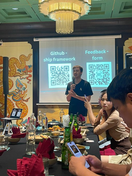 A man with a microphone stands before a projection screen displaying QR codes for 'Github - ship framework' and 'Feedback - form' at what appears to be a formal dinner or conference event.