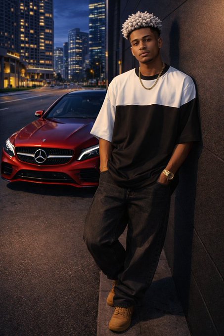 A young man with curly hair, wearing a black and white t-shirt, gold chain, and Timberland boots, leans against a wall at night with a red Mercedes-Benz parked on the street behind him.