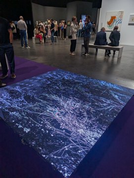 The image shows a group of people standing around an art exhibit, with some sitting on benches nearby. The floor has a unique design that appears to be made from purple tiles or a similar material, giving the impression of an endless garden growing on the ground. There are also several paintings displayed in the room, adding to the artistic atmosphere. The post accompanying this image provides information about the art exhibit and its location at Art Basel.