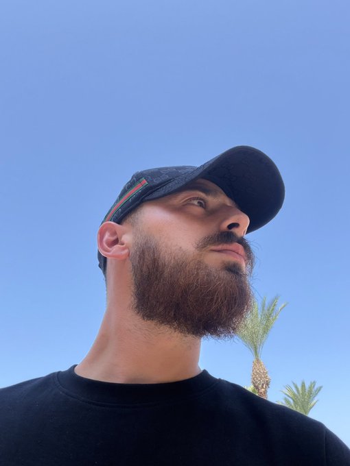 A bearded man wearing a black cap gazes upward against a clear blue sky with palm trees, projecting a confident and contemplative mood that aligns with the post's call to share Solana addresses for blessing three winners.