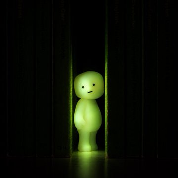 A small green glowing figure with a sad expression stands alone in a dark, narrow passage between black walls.