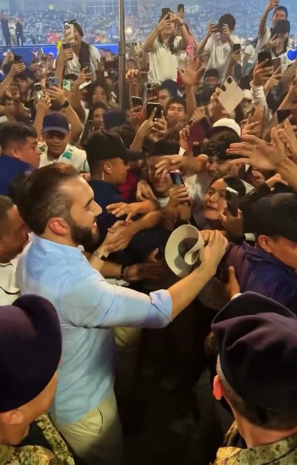 A bearded man in a light blue shirt greeting an enthusiastic crowd of fans holding up smartphones to take photos and videos at what appears to be an event.