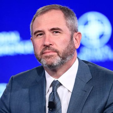 Brad Garlinghouse, CEO of Ripple, is shown in a blue suit and tie at what appears to be a formal conference, urging the Senate to pass the CLARITY Act. His serious expression reflects his firm stance that the legislation represents substantive policy work rather than a watered-down compromise.