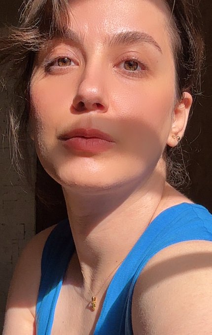 A close-up selfie of a woman with dark hair, wearing a blue tank top and small gold earrings, illuminated by bright sunlight with a shadow cast across her face.