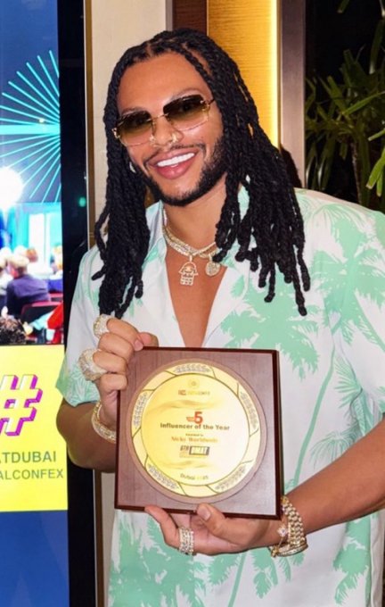 A smiling man with long dreadlocks, sunglasses, and jewelry holds a 'Influencer of the Year' award plaque at what looks like the Dubai Influencer Confex.