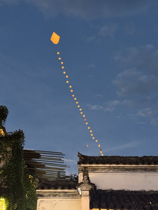 A glowing orange sky lantern connected to a long string of smaller lights floats against a blue evening sky above traditional Chinese rooftops with tiled eaves.