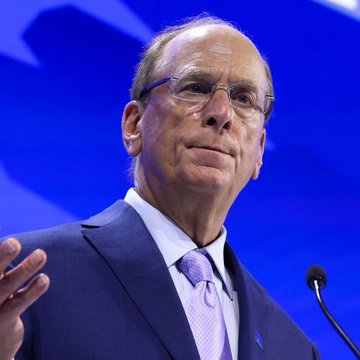 Larry Fink, CEO of BlackRock, speaks at a podium with a microphone, gesturing with his hand against a blue background. The image relates to the post as BlackRock—the firm he leads—has clients purchasing over $111 million worth of Bitcoin ($BTC).