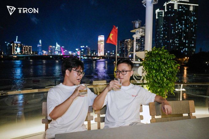 Two young men in white t-shirts sitting on a rooftop or balcony at night with a Vietnamese flag and illuminated city skyline behind them, with a 'TRON' logo in the top left corner.
