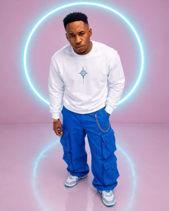 A man poses in a white long-sleeve shirt with a small cross logo and baggy blue cargo pants with a chain accessory, standing in front of a glowing blue neon circle on a pink background.