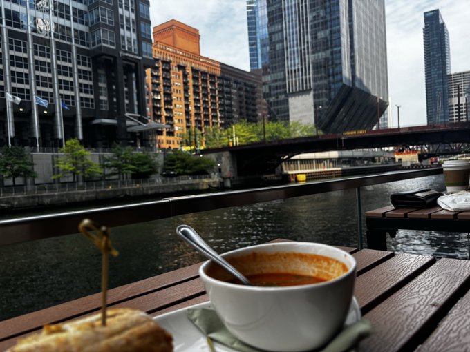 A bowl of orange soup on a wooden deck overlooking a river in an urban setting, with city buildings including modern skyscrapers and a bridge in the background.