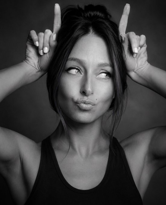 A black-and-white portrait of a woman looking to the side with pursed lips, holding her hands up with index fingers pointing outward near her head.