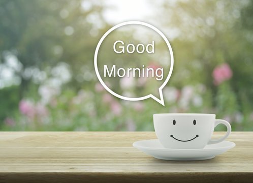 A cheerful morning greeting image featuring a white smiley-face coffee cup on a saucer on a wooden table, with a speech bubble saying 'Good Morning' against a soft, blurred garden background.