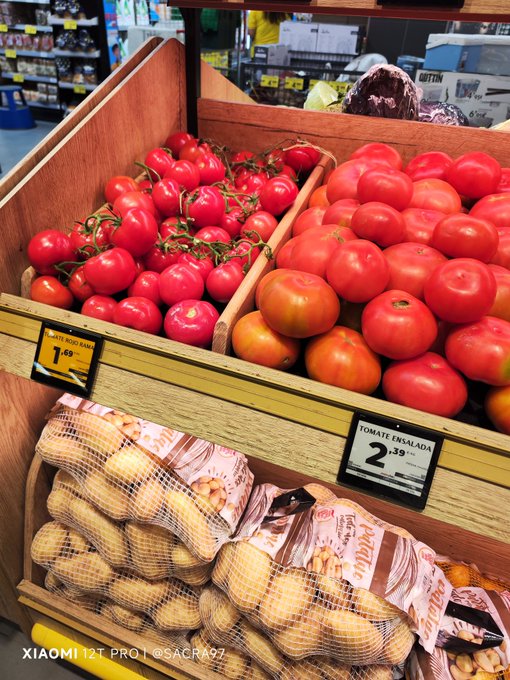 The image features a display of fresh vegetables, specifically potatoes, onions, and tomatoes, neatly arranged at a store. There are several bags of potatoes available for sale, with two prominently displayed in front of the rest. The arrangement also includes an assortment of onions of different sizes, and a selection of ripe, red tomatoes placed alongside them. In addition to these vegetables, there is a price sign indicating that they cost 1.00 units each.