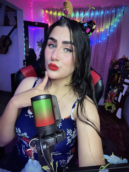 A woman with dark hair and red lipstick sits in a gaming chair behind a colorful RGB microphone, with string lights and plush toys in the background.