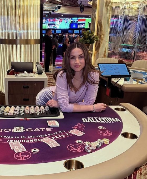 A young woman with long brown hair sitting at a casino blackjack table with the text 'BALLERINA' and 'NO GATE' visible on the felt.