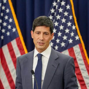A man in a dark suit and blue tie speaking at a podium, with several United States flags visible in the background behind him.