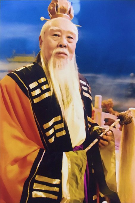 An actor in traditional Chinese costume portraying the elderly sage Jiang Ziya, with a long white beard, ornate yellow and black robes featuring I Ching trigrams, and a golden headdress.