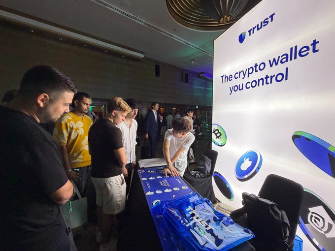 The image shows attendees gathered at a Trust Wallet booth featuring prominent signage reading "TRUST - The crypto wallet you control," with team members engaging visitors over a display table showcasing branded merchandise and informational materials at what appears to be a Hyperliquid community event in Singapore, illustrating the post's description of Trust Wallet's outreach and user engagement at the conference.