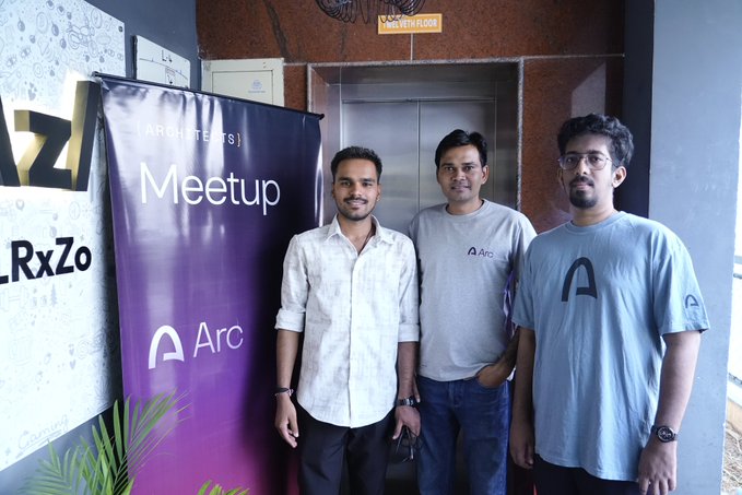 Three men pose for a photo at a tech Meetup event in front of a purple 'Architects Meetup' banner with the Arc browser logo, standing near an elevator lobby.