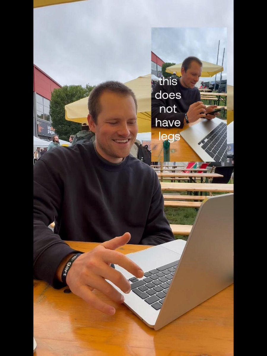 The image features a man sitting at an outdoor table with his laptop open in front of him. The scene takes place outdoors under umbrellas, suggesting that they might be enjoying their time outside while working or browsing the internet. There are also other people present in the background, indicating that this could be a popular spot for outdoor activities and socializing.