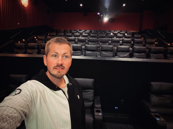 A man takes a selfie inside a dimly lit movie theater with rows of empty black reclining seats behind him.